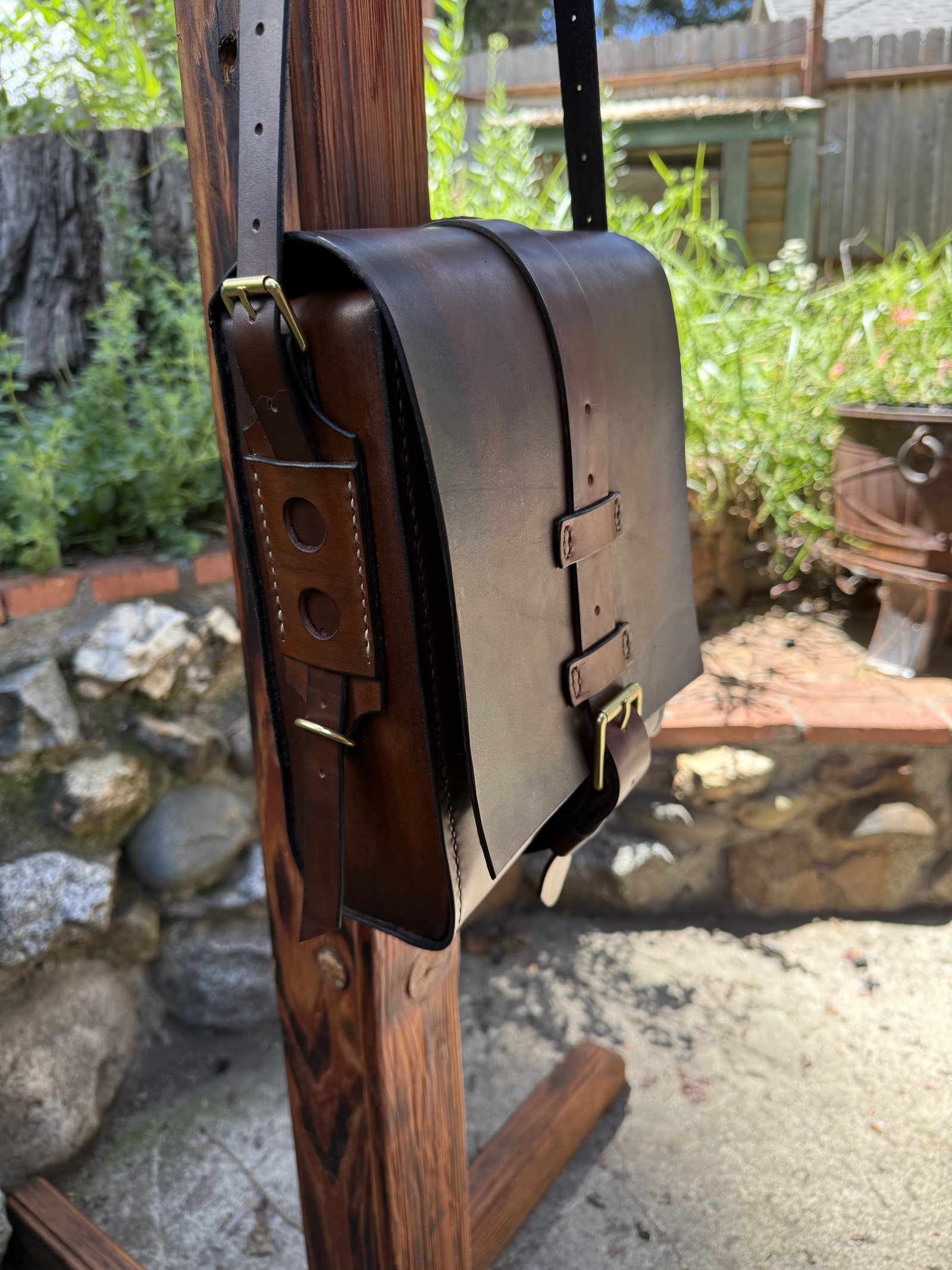 A dark brown flap-over satchel hanging from a wooden stand, closed with two strap-and-buckle fastenings in brass