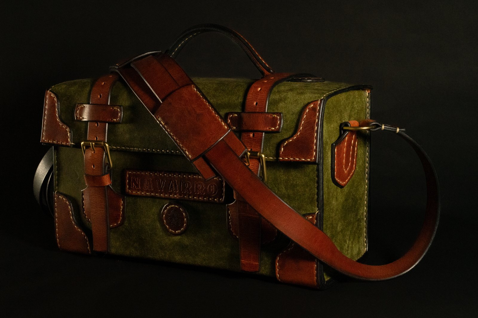 A range bag in olive-green suede and tan leather, photographed against black, its flap buckled shut over a name plate tooled to order, with the long shoulder strap laid across the front