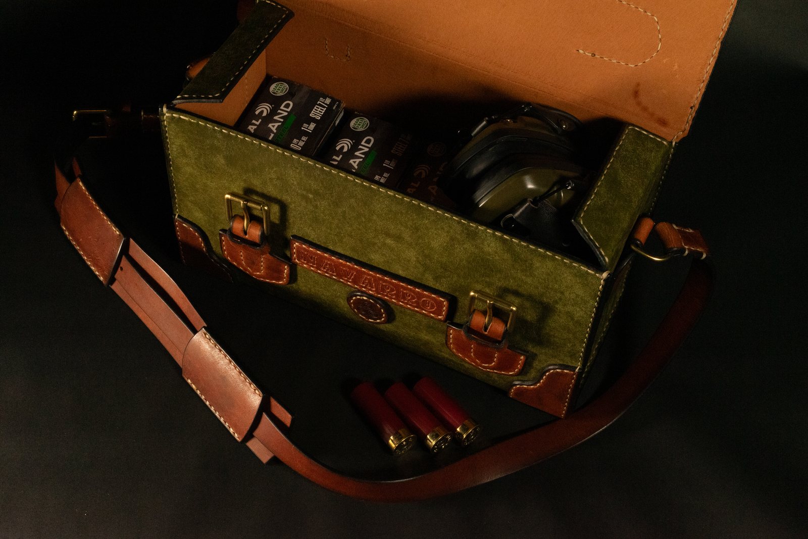The same range bag open, fitted out with boxes of shotshells and a set of ear defenders, three shells resting on the dark surface beside it