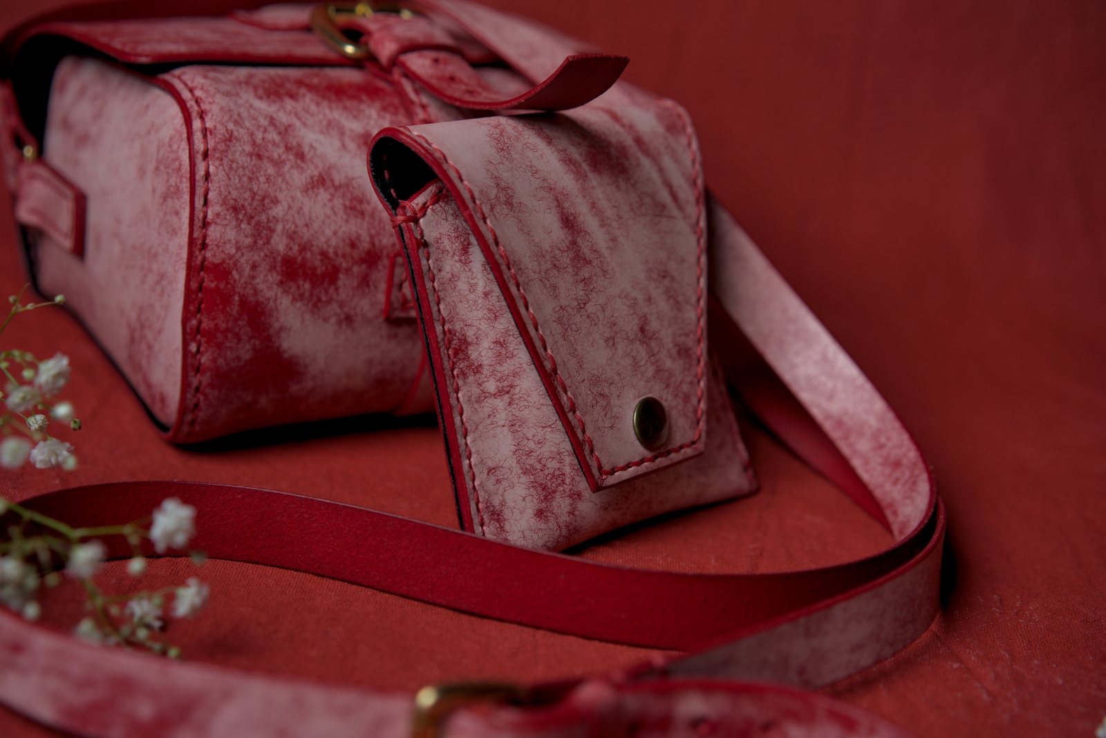 A boxy shoulder bag in red leather with a pale, mottled surface, a snap-fastened pocket on the front and its long strap looped across a red backdrop