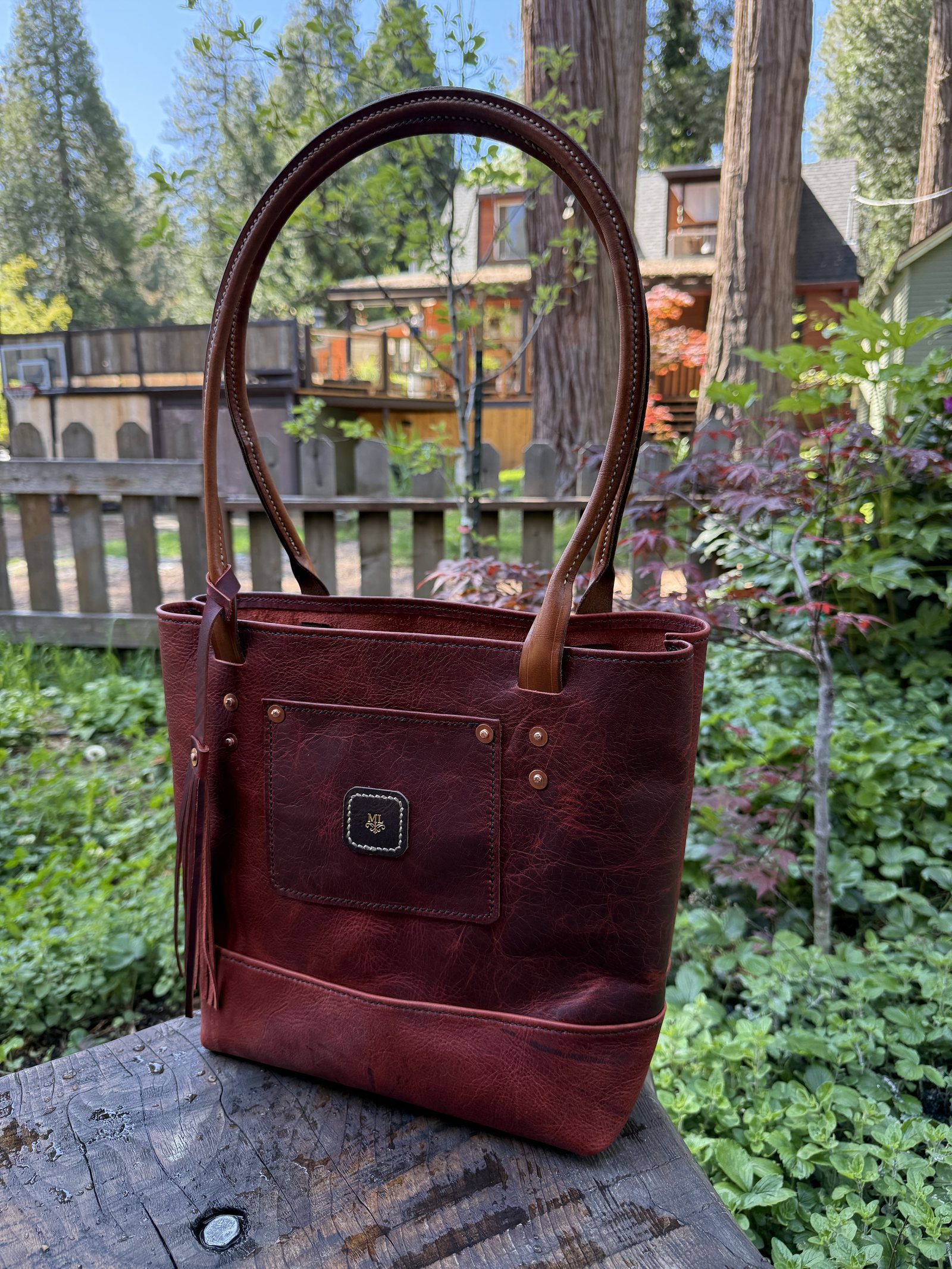 A deep red-brown leather tote standing on a stump, tan rolled handles, a riveted front pocket carrying the workshop's round mark, and a leather tassel at one side